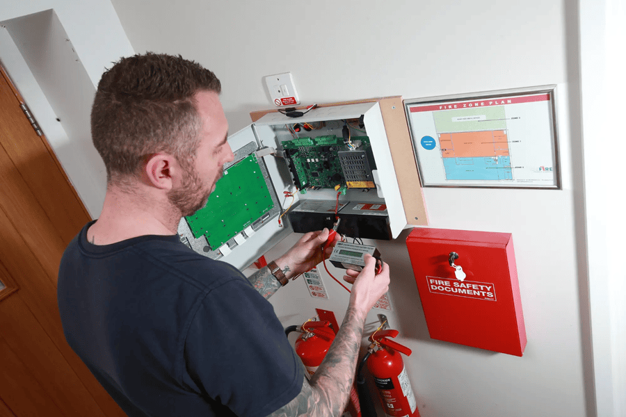 Fire Alarm Systems engineer testing a fire alarm control panel, with a fire zone plan and fire safety documents box on the wall alongside