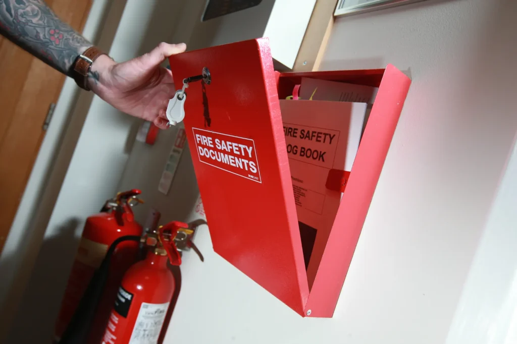 Hand opening a red fire safety documents cabinet containing a fire safety log book, with fire extinguishers alongside