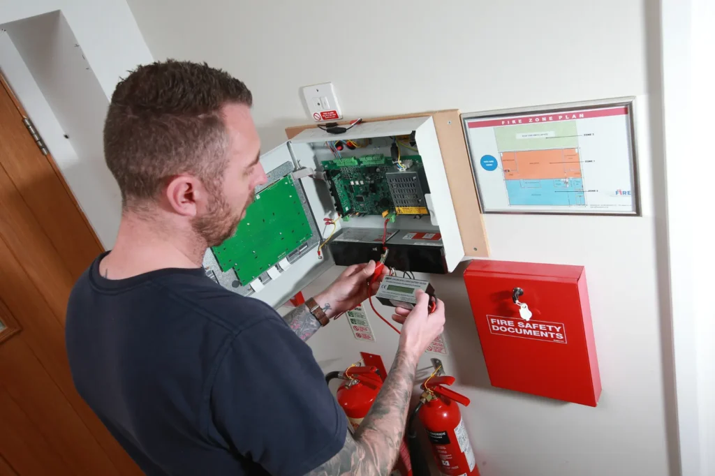 Fire Alarm Systems engineer testing the standby batteries inside an open fire alarm control panel, beside a wall-mounted fire zone plan