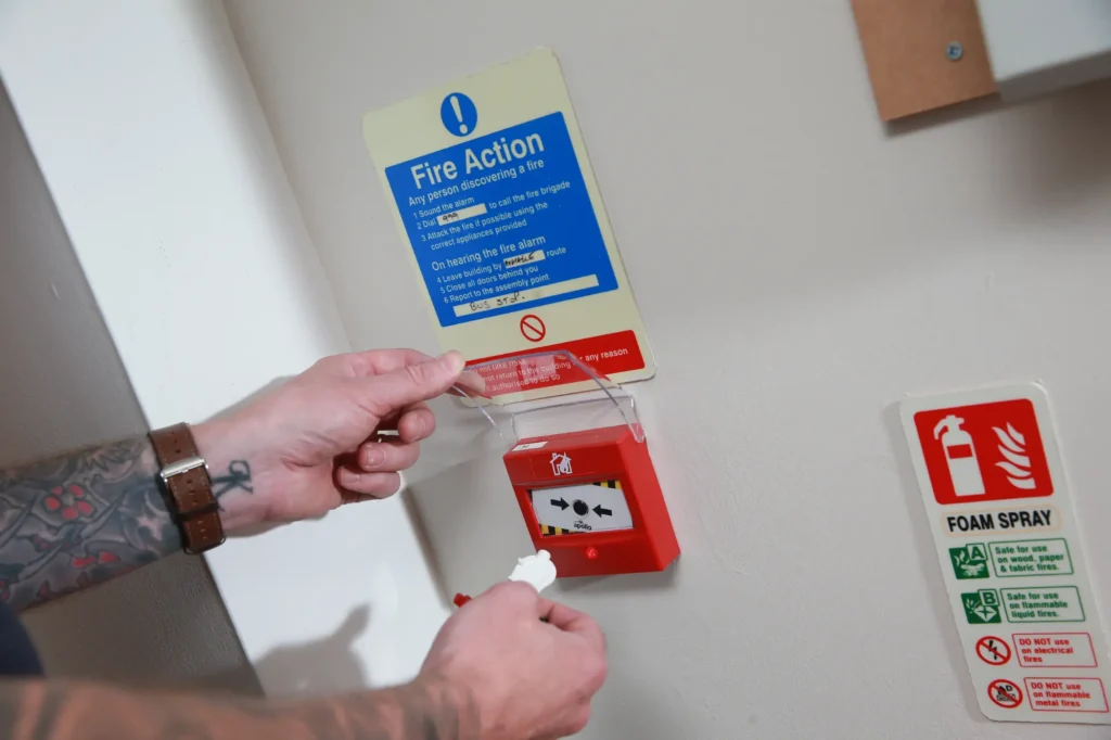 Fire Alarm Systems engineer testing an Apollo manual call point with a test key, below a Fire Action notice