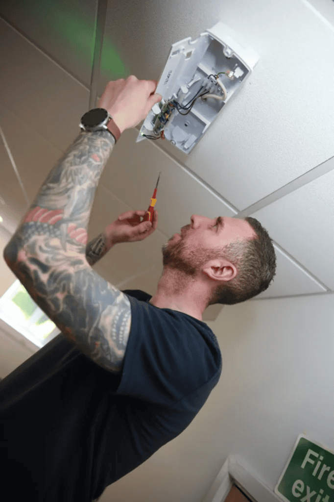 Fire Alarm Systems engineer using a screwdriver to service a ceiling-mounted emergency light fitting, with a fire exit sign nearby